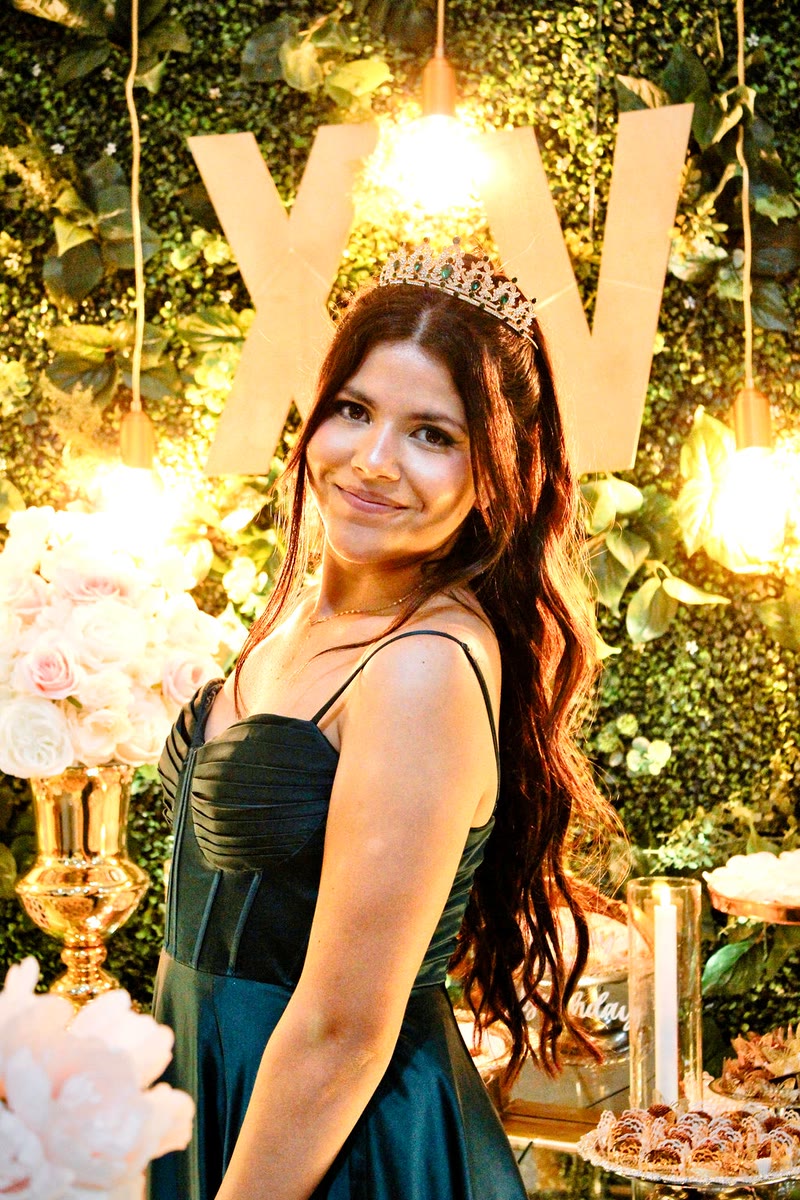 Girl in a dark green dress and tiara smiling in front of a floral backdrop with gold decorations